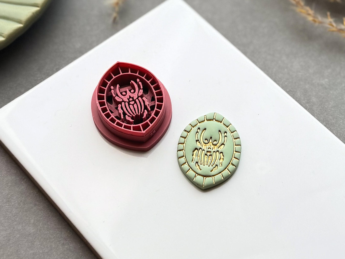 Goliath beetle oval frame polymer clay cutter displayed beside a matching finished polymer clay embellishment featuring a detailed Goliath beetle within a decorative geometric border.