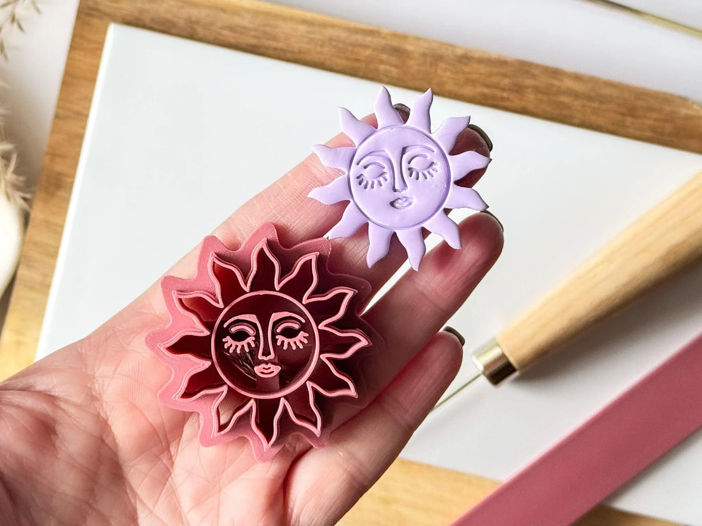 Hand holding a pink celestial sun polymer clay cutter with a matching lavender clay sun face cut-out for scale.