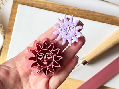 Hand holding a pink celestial sun polymer clay cutter with a matching lavender clay sun face cut-out for scale.