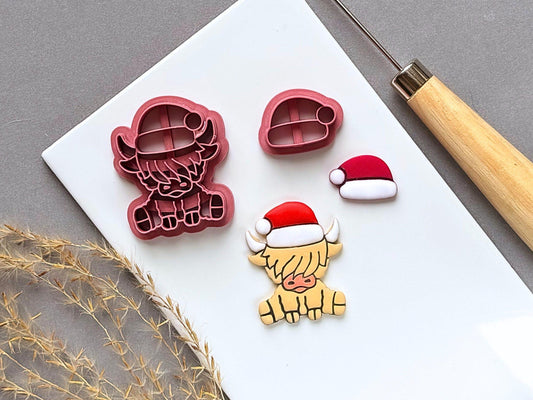 Highland cow Christmas polymer clay cutter featuring a Santa hat, fluffy fringe, horns and hooves, displayed alongside a separate Santa hat cutter and matching finished clay embellishments on a white tile.