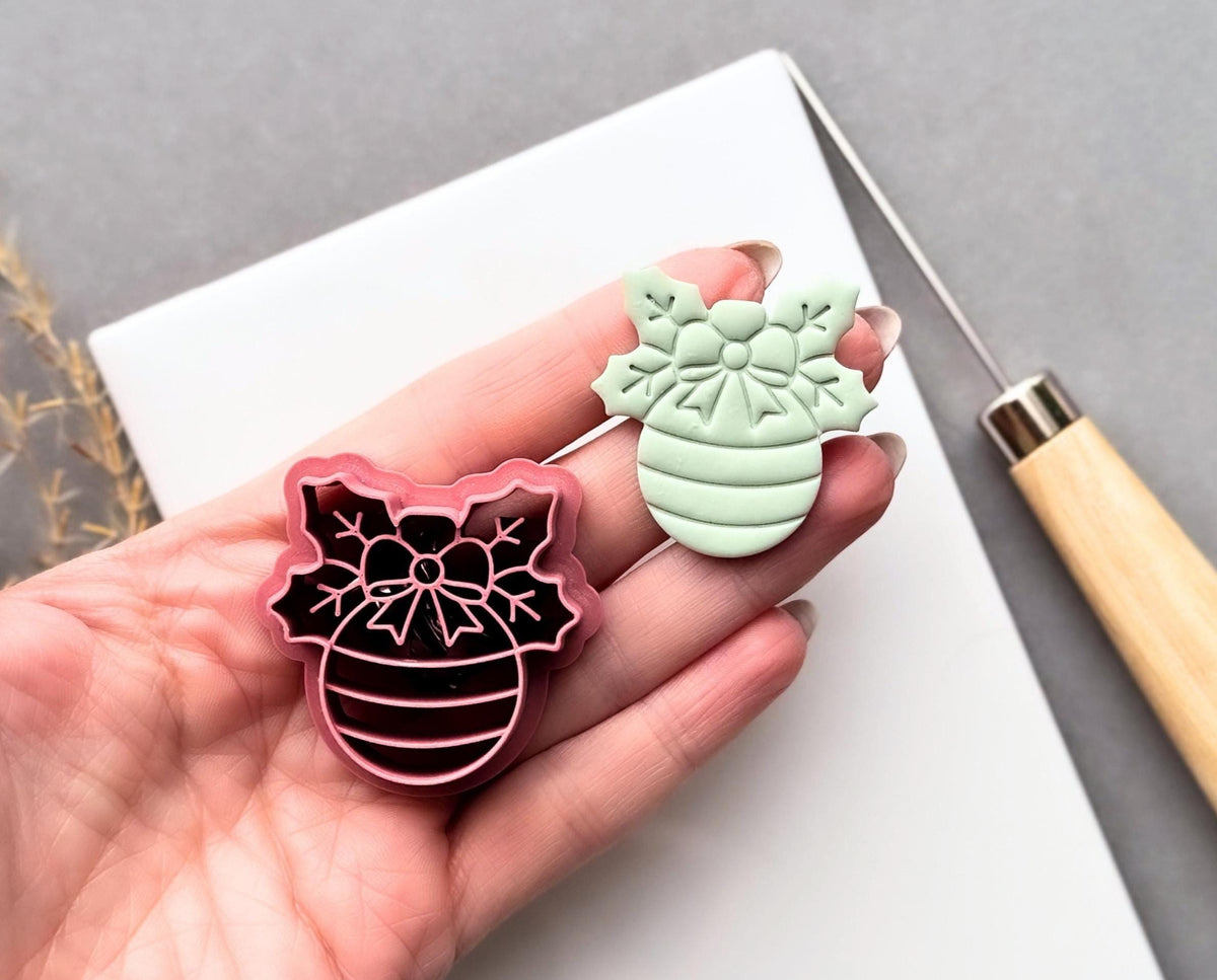 Christmas bauble polymer clay cutter held in a hand beside a matching finished clay cut-out, featuring a decorative snowflake bow, floral centre and striped bauble design.