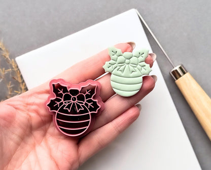 Christmas bauble polymer clay cutter held in a hand beside a matching finished clay cut-out, featuring a decorative snowflake bow, floral centre and striped bauble design.