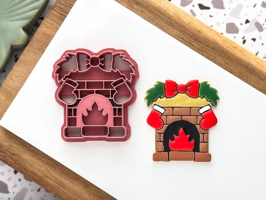 Festive brick fireplace clay cutter with burning fire, hanging stockings, bow, and leaf decorations.