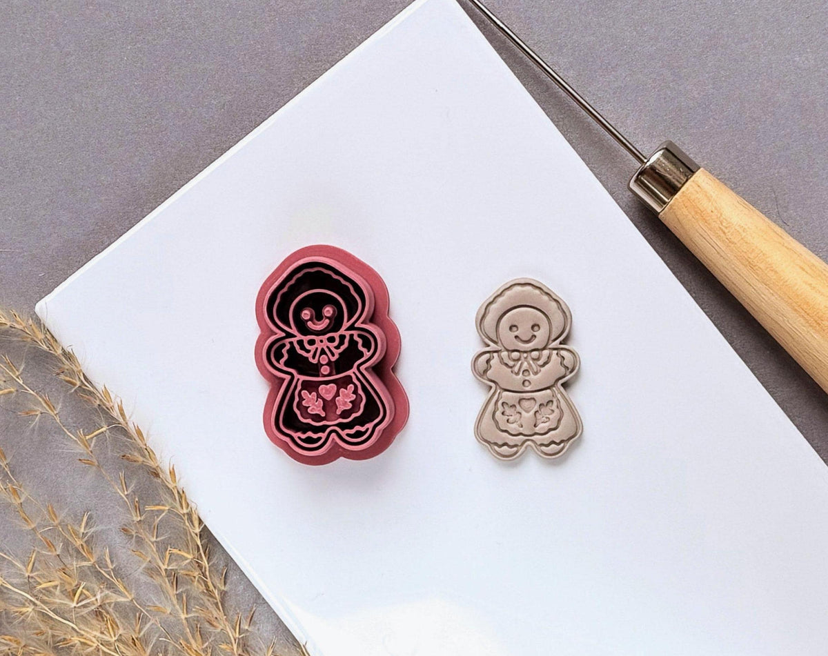 Vintage-inspired gingerbread girl polymer clay cutter featuring a bonnet, decorative collar, smiling face and folk-art floral apron details, displayed beside a matching finished clay embellishment on a white tile.