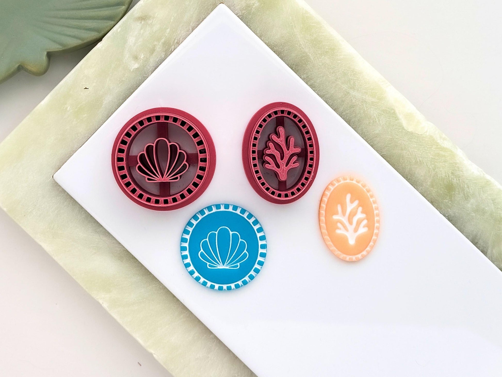 Pink coastal clay cutters featuring a clam shell cutter and coral clay cutter, displayed with blue and pastel orange summer clay sample pieces on a white tile with a green marble background.