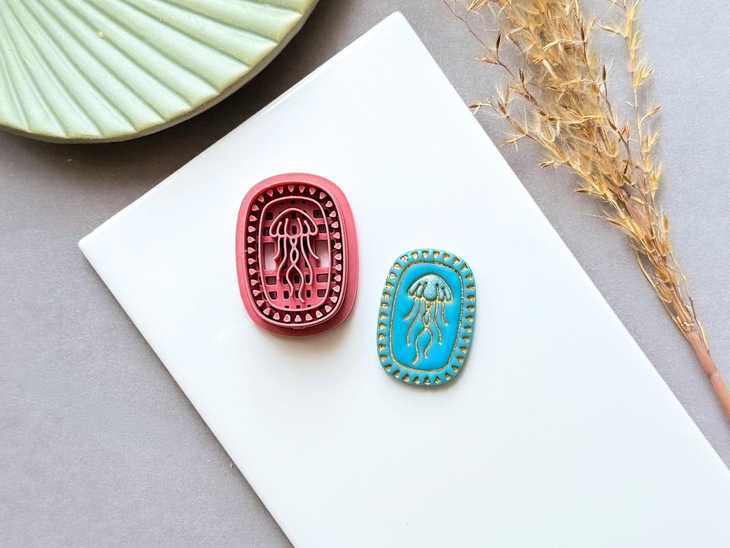 Jellyfish polymer clay cutter displayed alongside a finished polymer clay jellyfish sample, featuring an oval frame with intricate jellyfish embossing for creating ocean-inspired jewellery, charms and coastal craft projects.