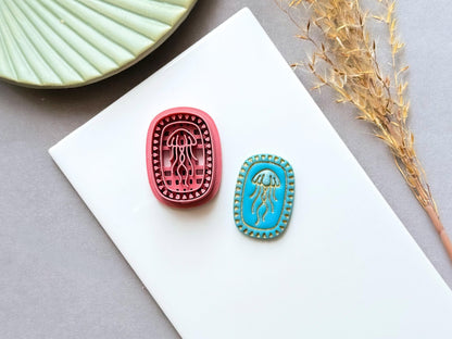 Jellyfish polymer clay cutter displayed alongside a finished polymer clay jellyfish sample, featuring an oval frame with intricate jellyfish embossing for creating ocean-inspired jewellery, charms and coastal craft projects.