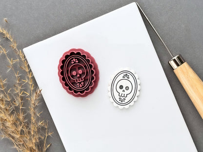 Gothic skull cameo polymer clay cutter displayed beside a matching finished clay cut-out, featuring a skull with decorative floral details inside an ornate scalloped oval frame.