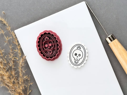 Gothic skull cameo polymer clay cutter displayed beside a matching finished clay cut-out, featuring a skull with decorative floral details inside an ornate scalloped oval frame.