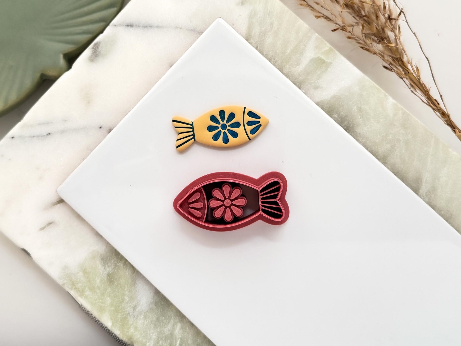 Finless fish shaped polymer clay cutter with eye, floral patterns and tail detail. The cutter is pink. Next to it is a beige clay cut out made with the cutter. It is used for making summer and ocean themed earrings.