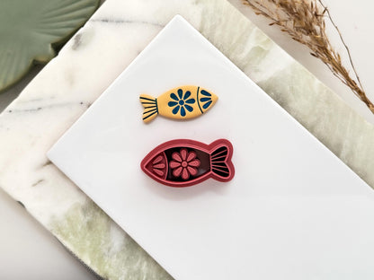 Finless fish shaped polymer clay cutter with eye, floral patterns and tail detail. The cutter is pink. Next to it is a beige clay cut out made with the cutter. It is used for making summer and ocean themed earrings.