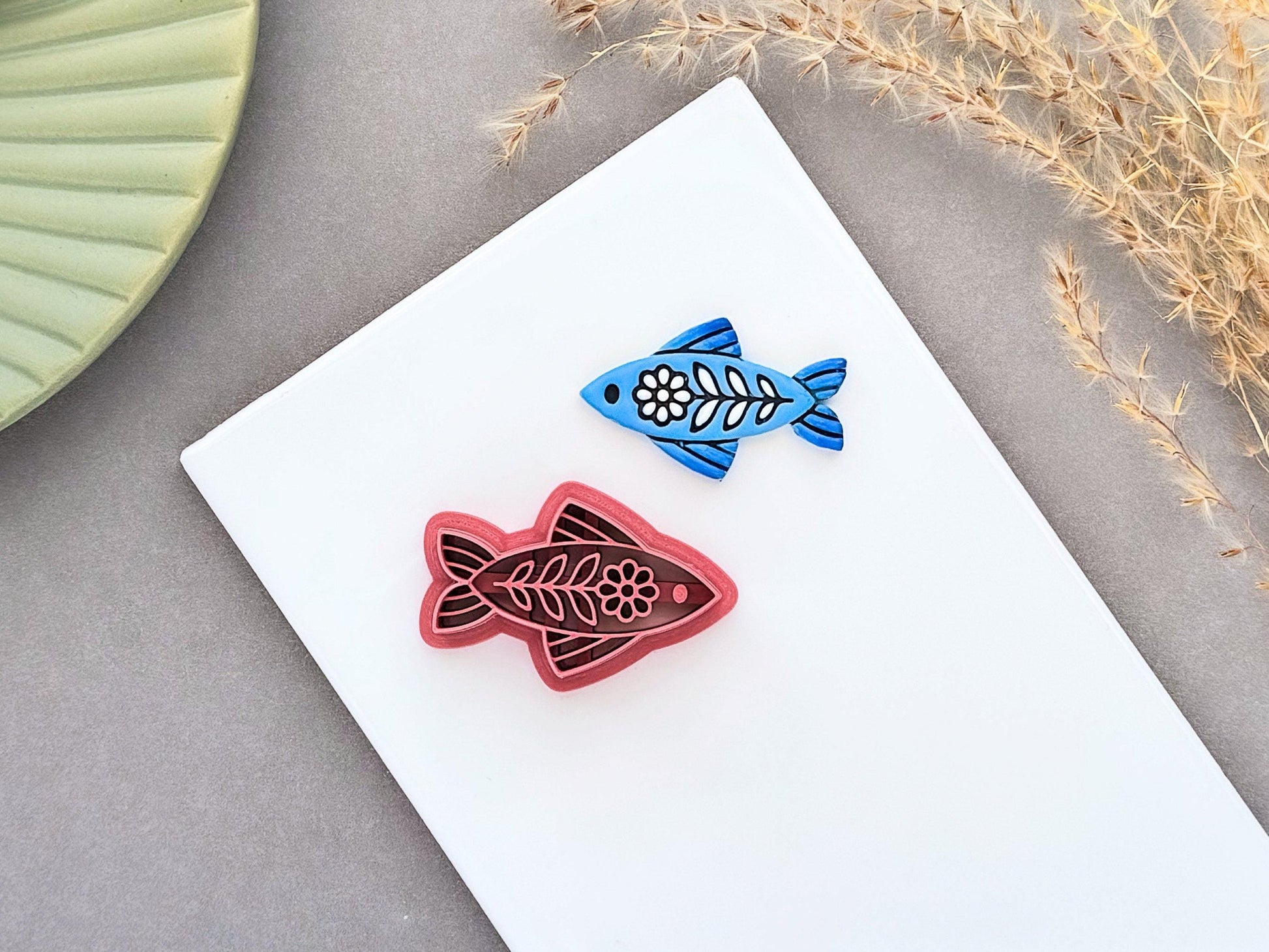 Blue folk art fish clay charm beside a dusty pink ornamental fish polymer clay cutter on a white ceramic tile.