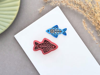 Blue folk art fish clay charm beside a dusty pink ornamental fish polymer clay cutter on a white ceramic tile.