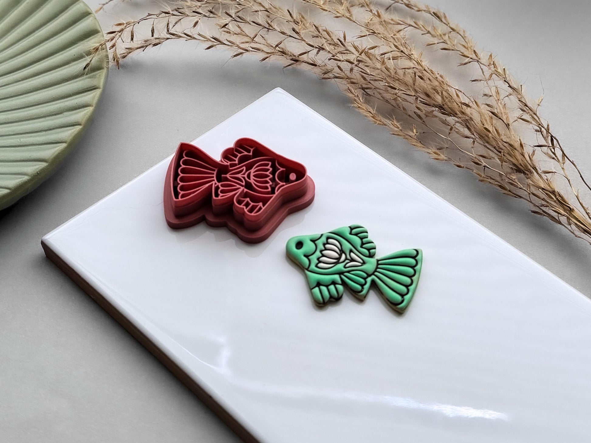 Side view of an ornamental fish polymer clay cutter displayed beside a matching mint green clay cut-out. The folk art-inspired fish design features decorative fins and floral details on a white ceramic tile.