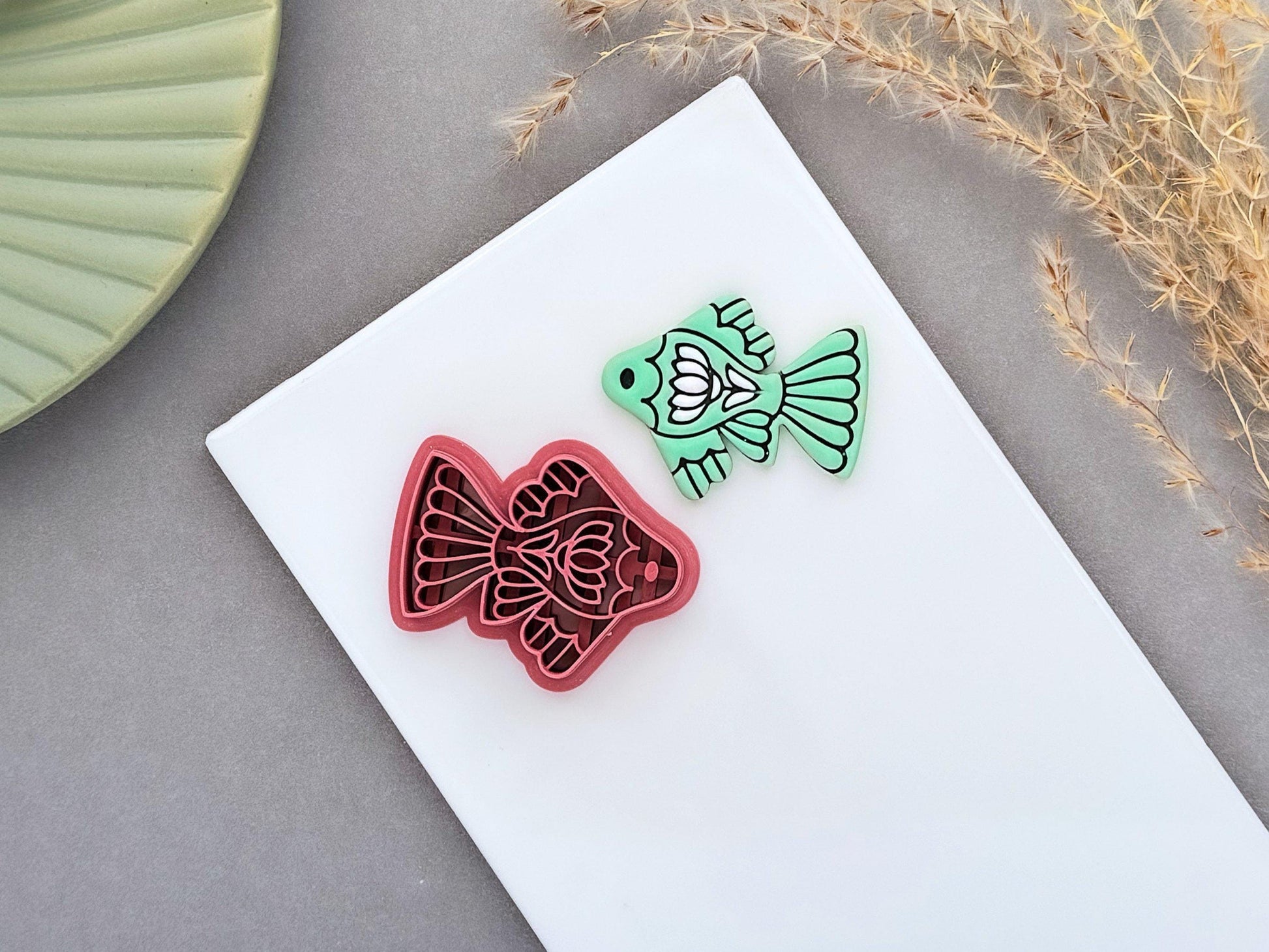 Ornamental fish polymer clay cutter displayed beside a matching mint green clay cut-out. The folk art-inspired fish design features decorative fins and floral details and is photographed on a white ceramic tile.