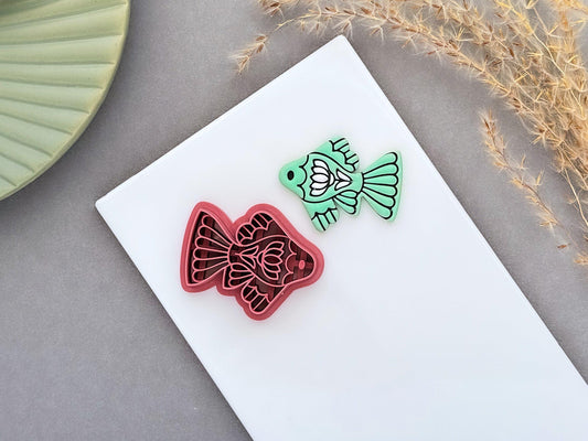 Ornamental fish polymer clay cutter displayed beside a matching mint green clay cut-out. The folk art-inspired fish design features decorative fins and floral details and is photographed on a white ceramic tile.