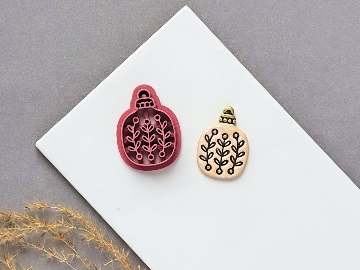 Floral sprig Christmas bauble polymer clay cutter displayed beside a matching finished clay embellishment featuring a symmetrical botanical leaf pattern.