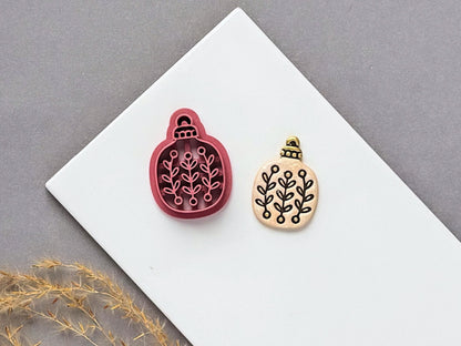 Floral sprig Christmas bauble polymer clay cutter displayed beside a matching finished clay embellishment featuring a symmetrical botanical leaf pattern.