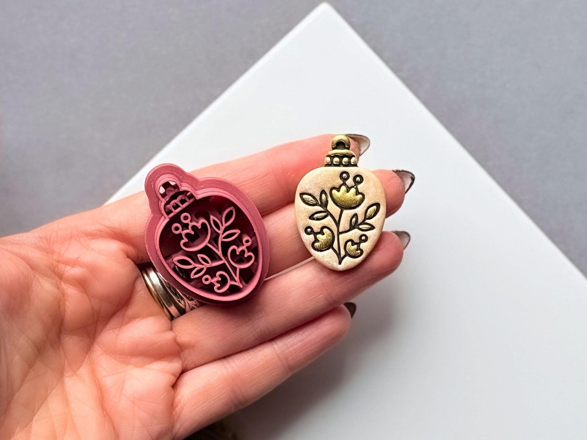 Folk art floral Christmas bauble polymer clay cutter held in a hand beside the matching finished clay cut-out, featuring a decorative flower, curling stems and leafy branches within an oval bauble shape.