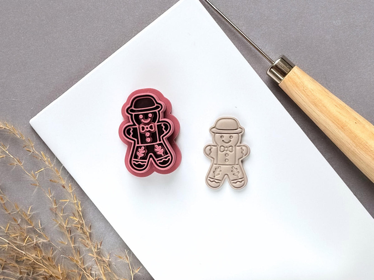 Vintage-inspired gingerbread man polymer clay cutter featuring a bowler hat, bow tie, buttons and folk-art floral leg details, displayed beside a matching finished clay embellishment on a white tile.