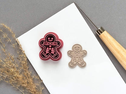 Gingerbread person polymer clay cutter featuring a smiling face, bow tie, buttons and decorative icing details, displayed beside a matching finished clay embellishment on a white tile.
