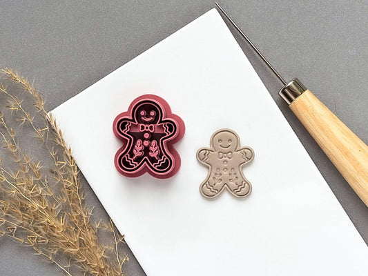 Gingerbread person polymer clay cutter featuring a smiling face, bow tie, buttons and decorative icing details, displayed beside a matching finished clay embellishment on a white tile.