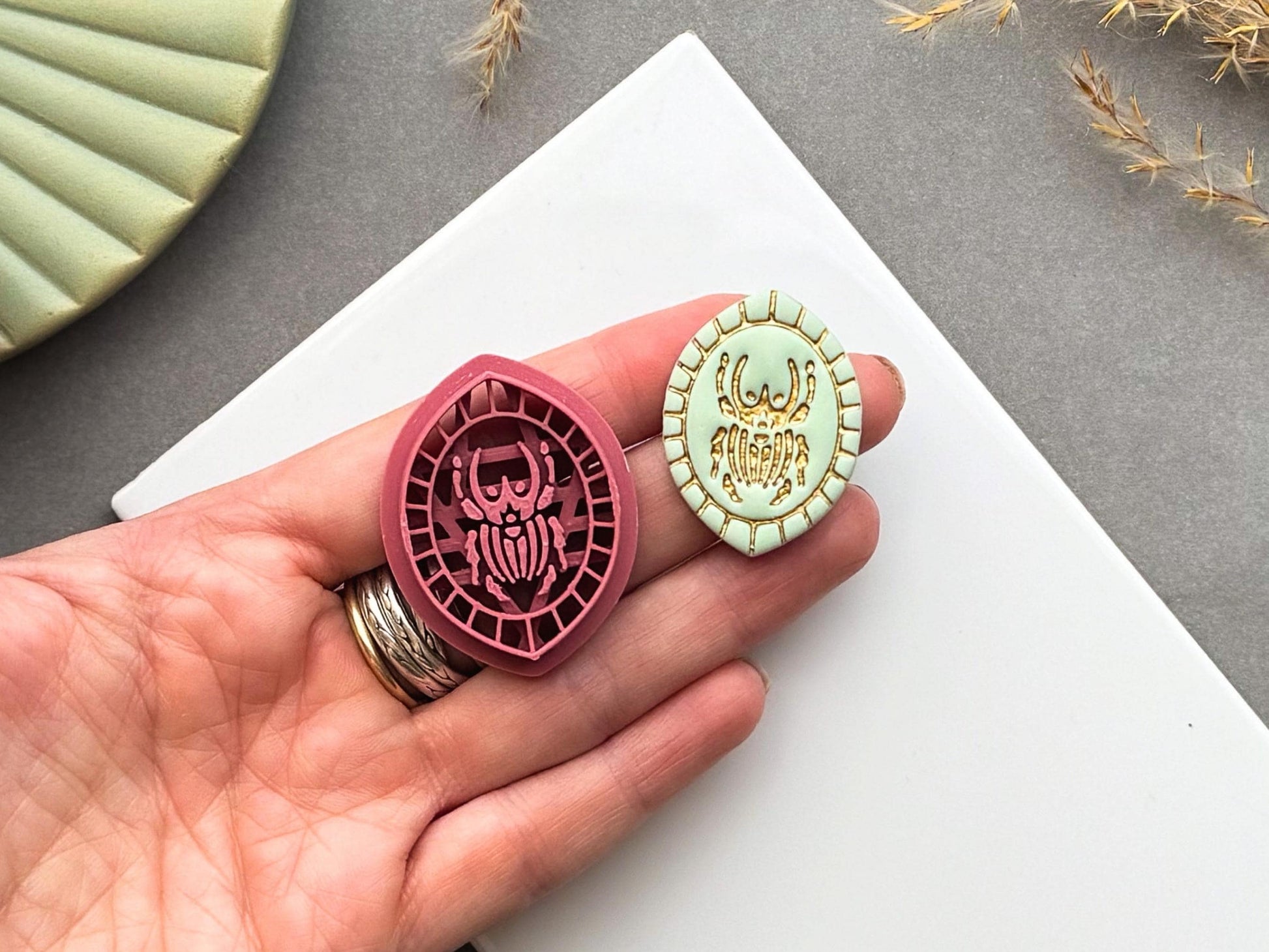 Goliath beetle polymer clay cutter and matching finished polymer clay embellishment held in a hand for scale, featuring a detailed Goliath beetle within a decorative geometric border.