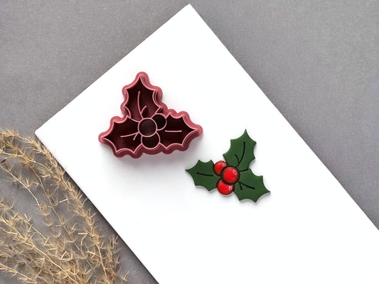 Christmas holly polymer clay cutter displayed beside a matching finished clay cut-out, featuring three berries surrounded by detailed holly leaves with pointed edges and vein imprints.