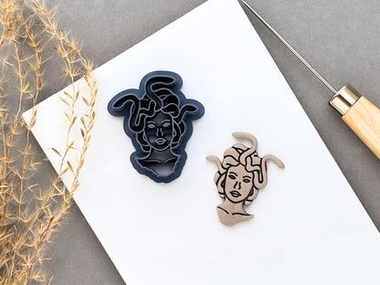 Medusa head clay cutter featuring a detailed face surrounded by flowing snakes, shown beside a finished cream polymer clay Medusa cut-out with black line details. Gothic and mythological-inspired design displayed on a white surface.