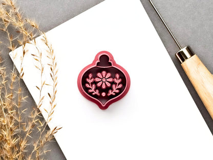 Folk art floral Christmas bauble clay cutter featuring a central flower, leafy botanical details and a hanging loop. Pink 3D-printed cutter displayed on a white surface.