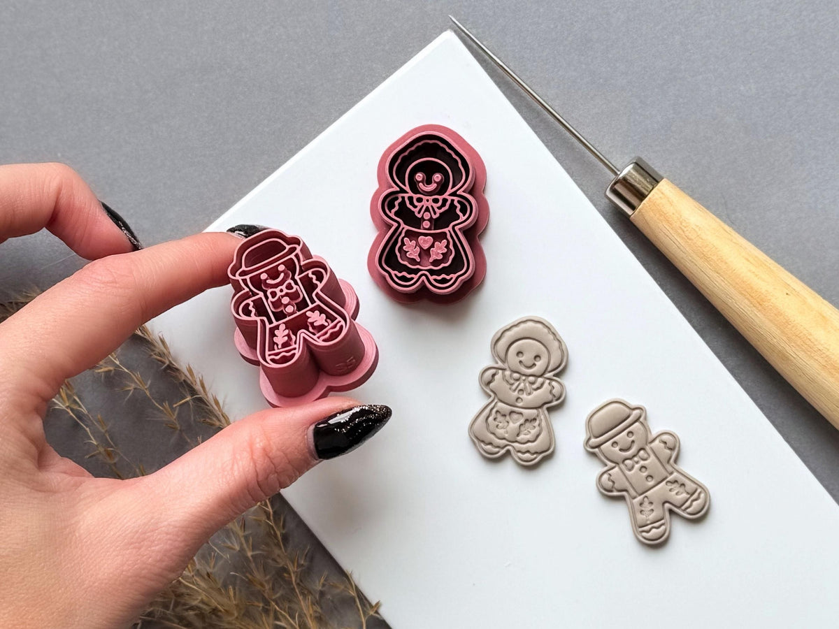Vintage-inspired gingerbread man and gingerbread girl polymer clay cutters, featuring folk-art floral motifs, decorative icing details, hats, bow ties and smiling faces. One cutter is held in a hand to showcase the raised stamp design, with matching finished clay embellishments displayed on a white tile.