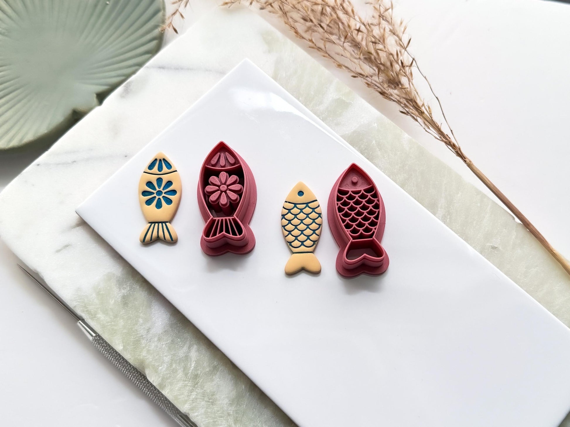 Dark pink coloured simple fish shaped clay cutters, one cutter has a folk style floral pattern, and the other has a scales pattern. Next to them are polymer clay sample cut outs made with the cutters and painted. All are on a white tile with decorative items.