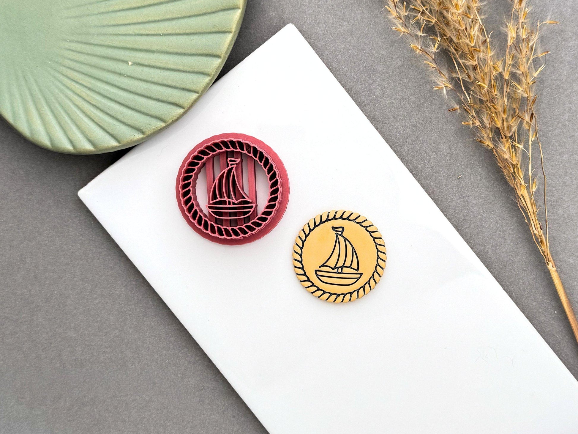 Round sailing boat polymer clay cutter beside the finished clay embellishment, featuring a rope border and detailed sailboat design for nautical themed creations.