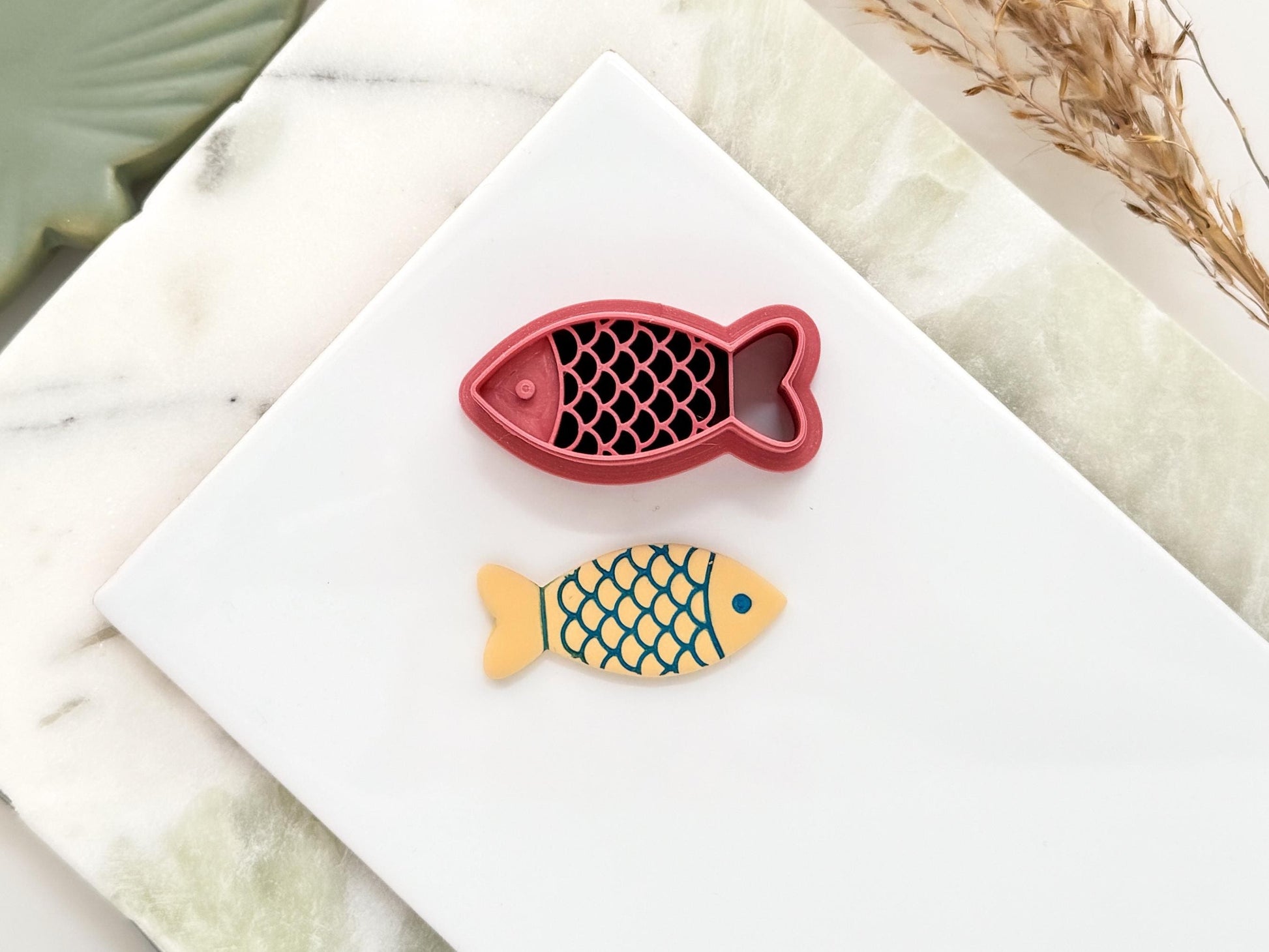 Finless fish shaped polymer clay cutter with eye, scales and tail detail. The cutter is pink. Next to it is a beige clay cut out made with the cutter. It is used for making summer and ocean themed earrings.