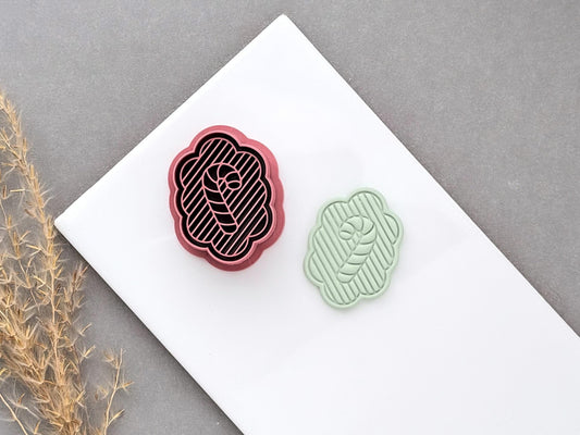 Christmas candy cane polymer clay cutter displayed beside a matching finished clay cut-out, featuring a scalloped frame, striped background and central candy cane design.