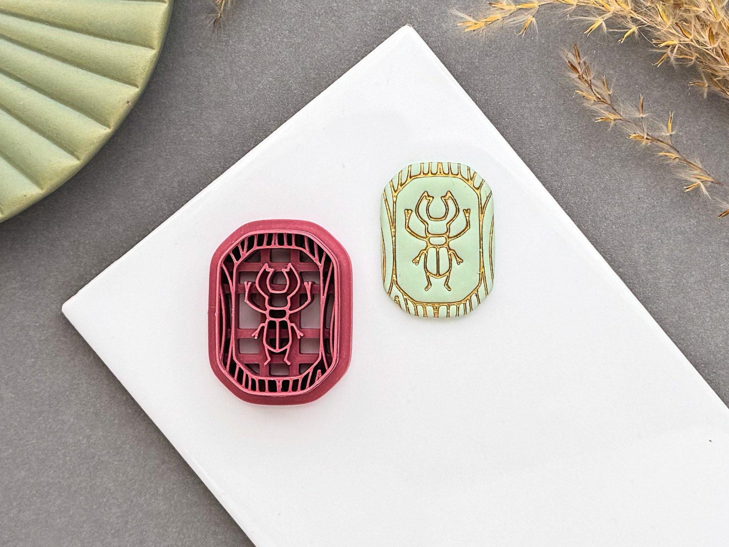 Scarab beetle polymer clay cutter displayed beside a matching finished polymer clay embellishment featuring a detailed scarab beetle within a decorative Egyptian-inspired border.