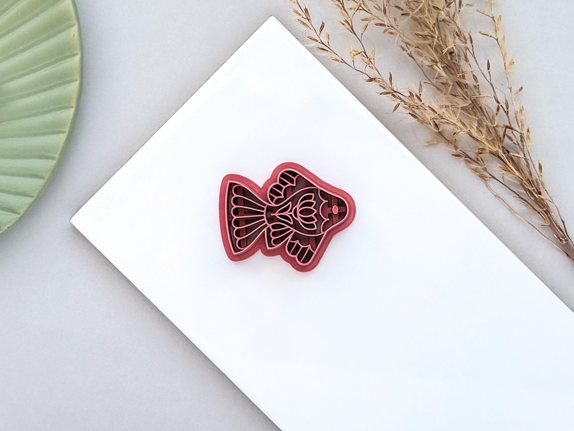 Dusty pink ornamental fish polymer clay cutter with folk art details displayed on a white ceramic tile with dried grasses.