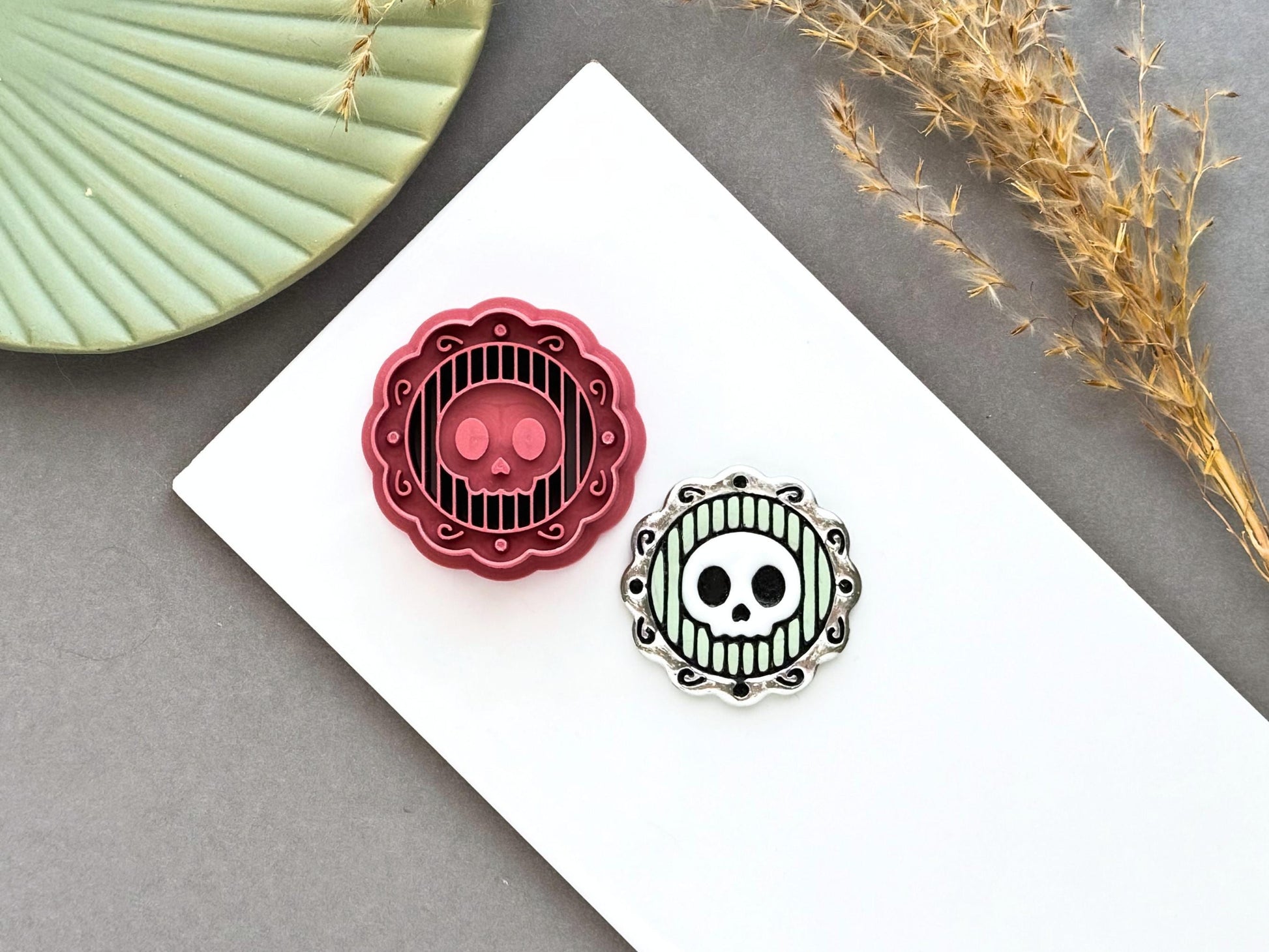 Skull cameo polymer clay cutter displayed alongside a finished polymer clay skull cameo sample, featuring a decorative ornate frame with striped background for creating gothic jewellery, Halloween charms and spooky craft projects.