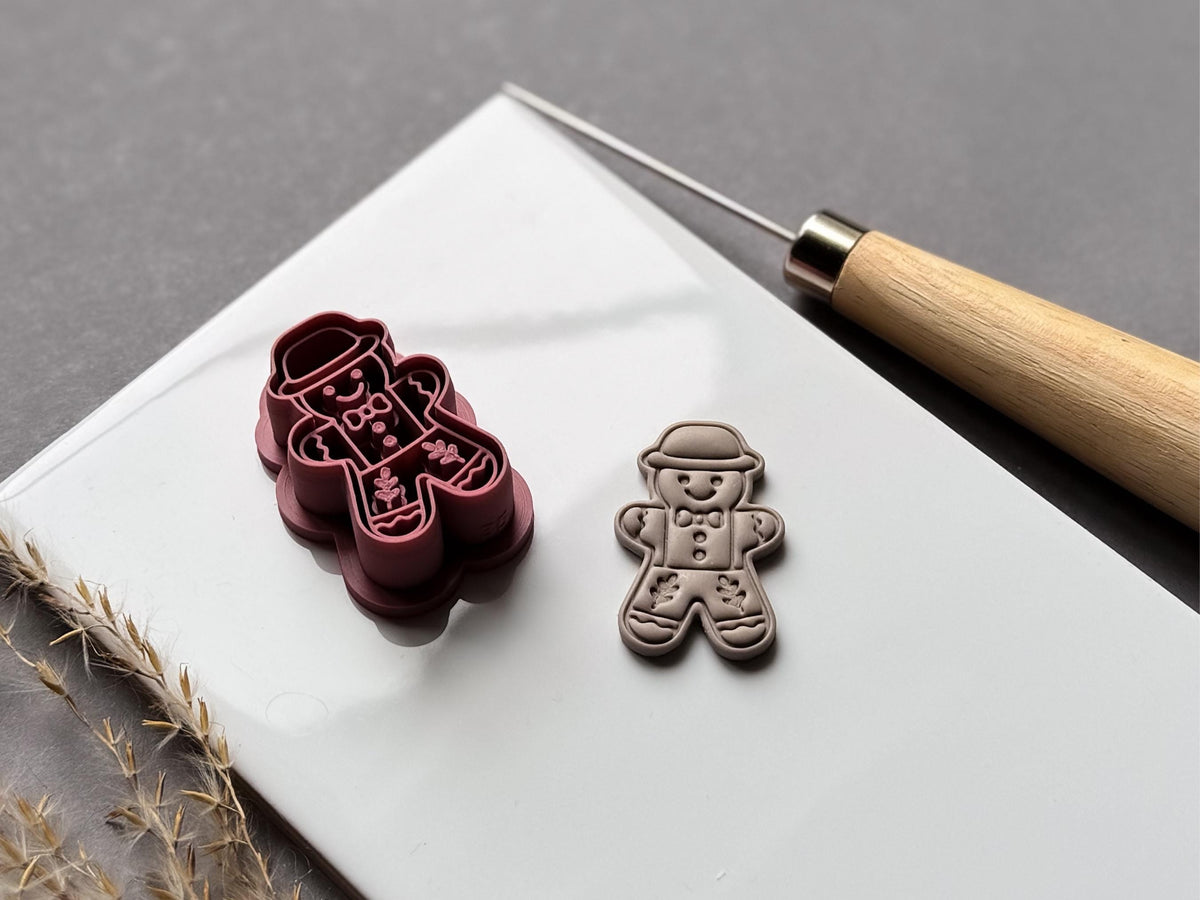 Vintage-inspired gingerbread man polymer clay cutter featuring a bowler hat, bow tie, buttons and folk-art floral leg details, displayed beside a matching finished clay embellishment on a white tile.