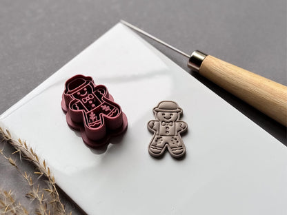 Vintage-inspired gingerbread man polymer clay cutter featuring a bowler hat, bow tie, buttons and folk-art floral leg details, displayed beside a matching finished clay embellishment on a white tile.