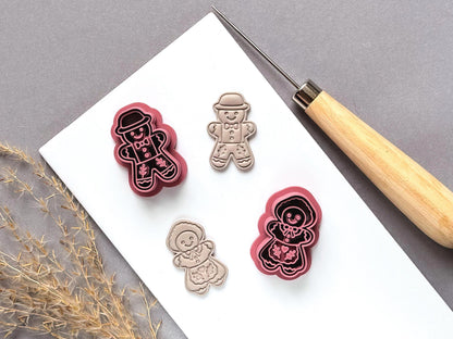 Vintage style gingerbread man and gingerbread girl polymer clay cutters displayed beside matching finished clay embellishments, featuring smiling faces, hats, bow ties, buttons and decorative icing details on a white tile.
