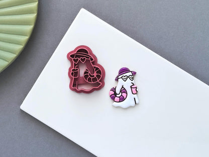 Ghost-shaped polymer clay cutter displayed beside a matching white clay cut-out. The summer-themed ghost design features a sun hat, heart-shaped sunglasses, a drink and a striped swim ring, photographed on a white ceramic tile.