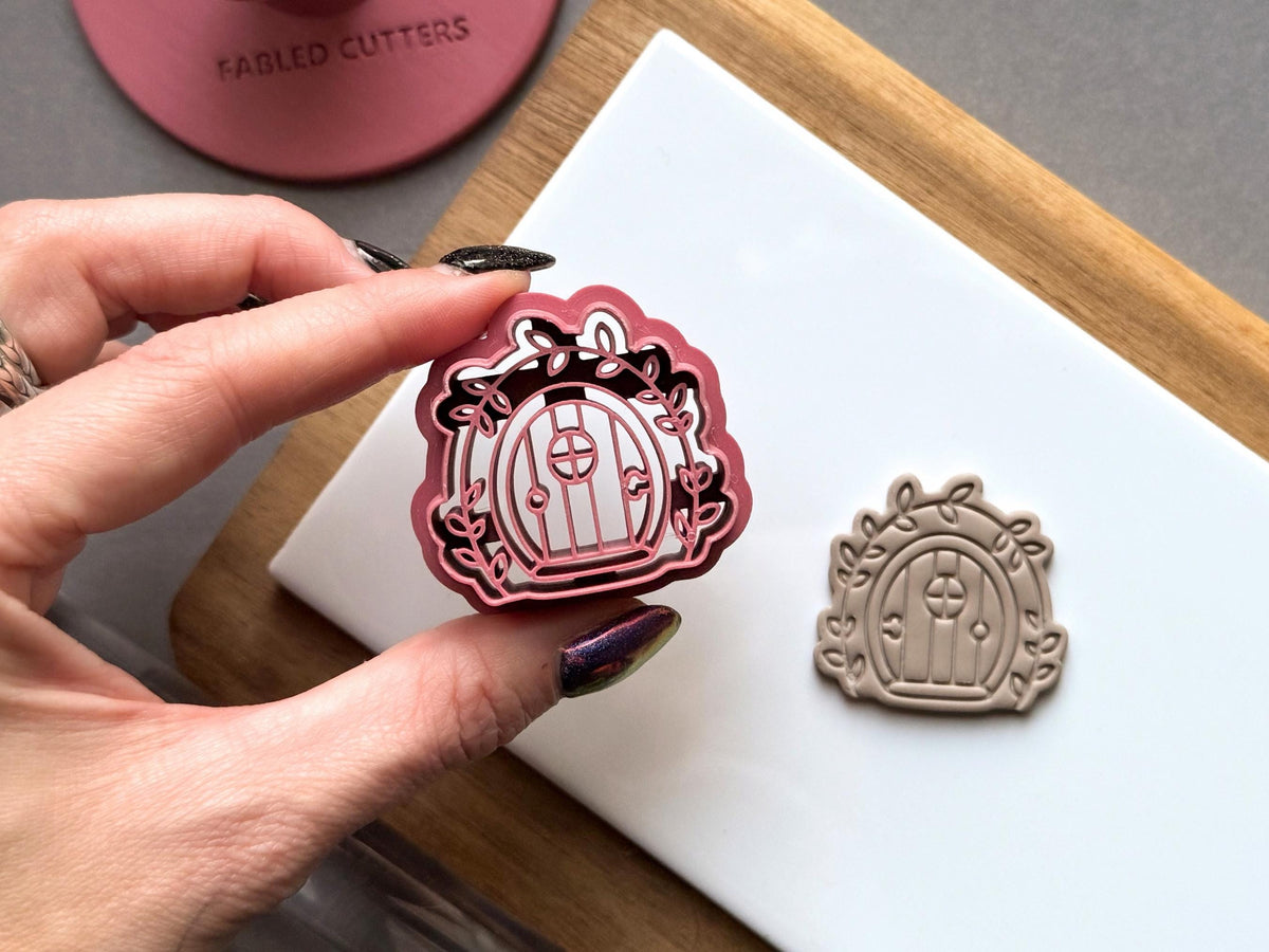 Fairy door polymer clay cutter held between fingers to showcase the arched door, round window, decorative leafy vines and scalloped outer frame, with a matching finished clay embellishment beside it.