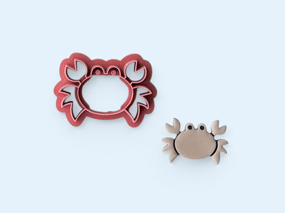 Pink crab shaped clay cutter with simple legs pincers and eyes next to a taupe polymer clay cut out crab. 