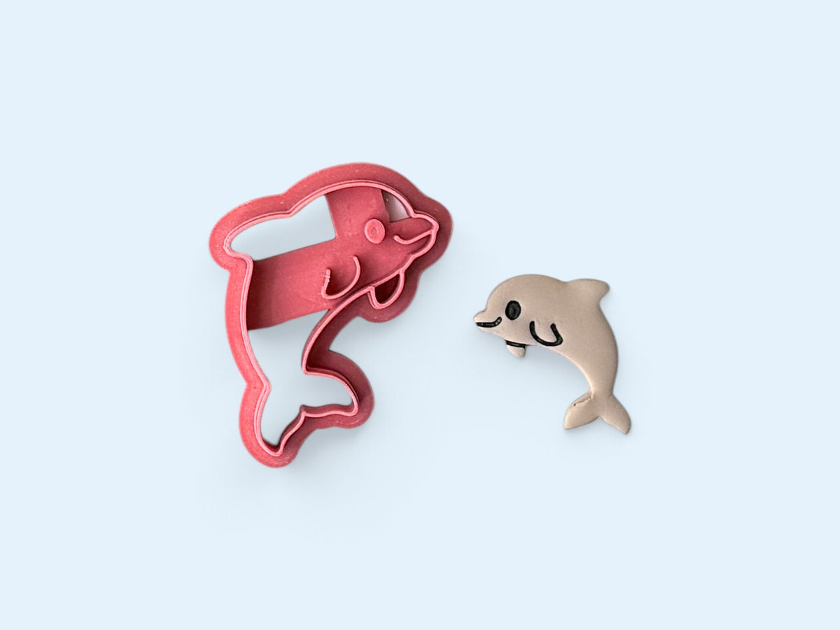 A pink dolphin-shaped polymer clay cutter shown next to a small beige polymer clay cutout of the same dolphin design on a light blue background.