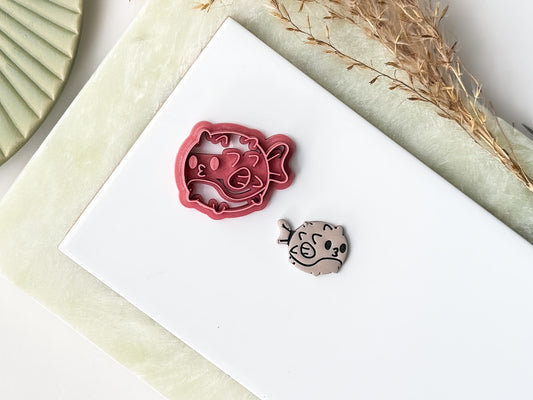 Pink spikey puffer fish shaped clay cutter with fin and face stamp details. Next to the cutter is a polymer clay cut out sample earring. Both are on a white baking tile.
