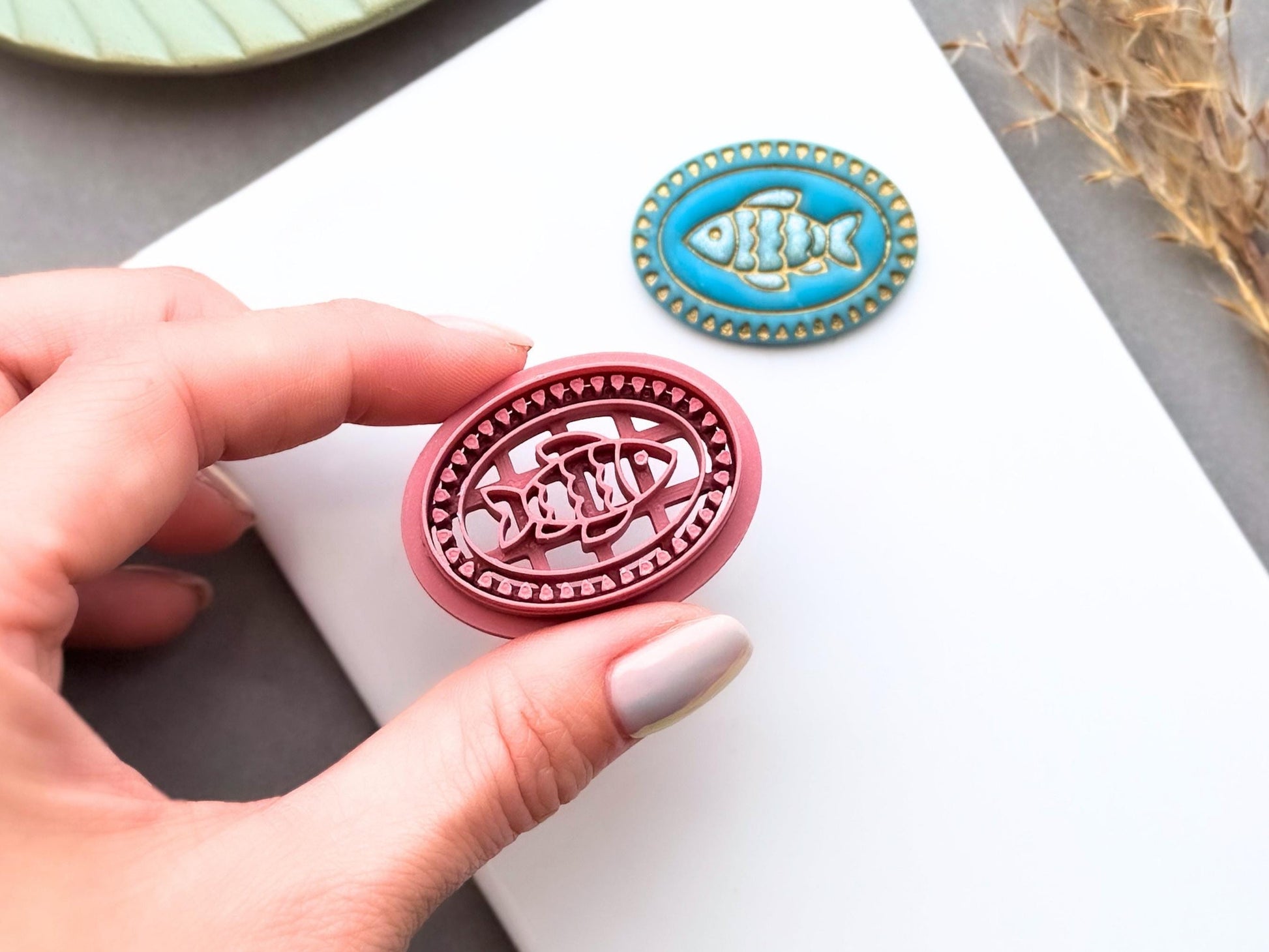 Fish polymer clay cutter shown being held in a hand to demonstrate scale and intricate detail, displayed alongside a finished polymer clay fish sample with a decorative beaded oval frame for creating coastal jewellery, ocean-inspired charms and marine craft projects.