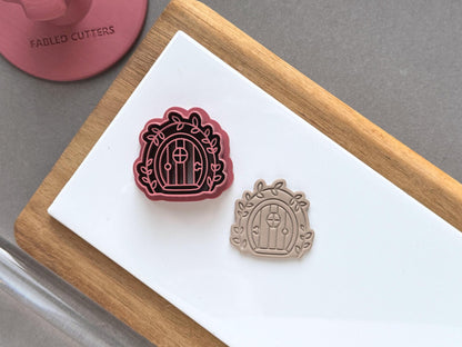 Fairy door polymer clay cutter displayed beside a matching finished clay embellishment, featuring an arched door with a round window, decorative leafy vines and a scalloped outer frame.