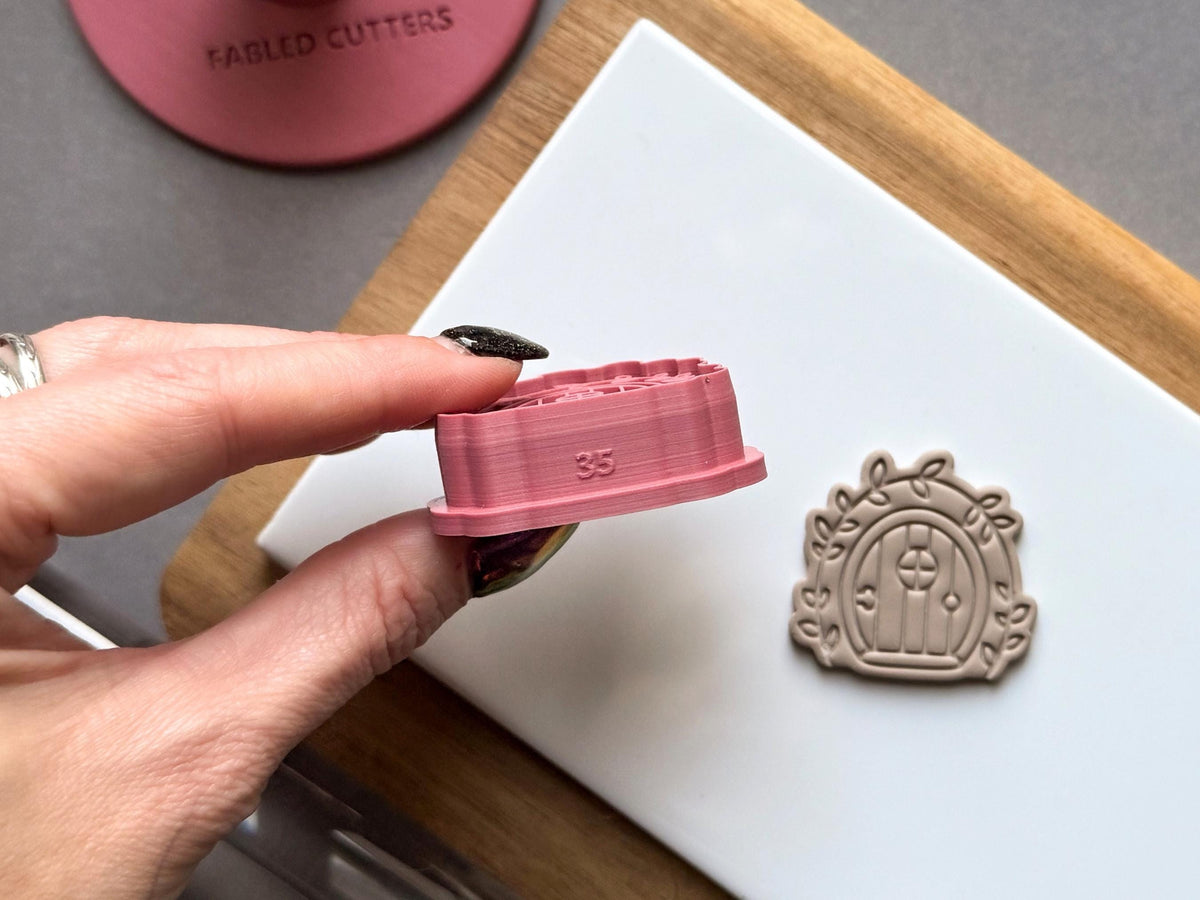 Fairy door polymer clay cutter held side-on to showcase the internal size marking and raised cutting wall, with a matching finished clay embellishment displayed on a white tile.