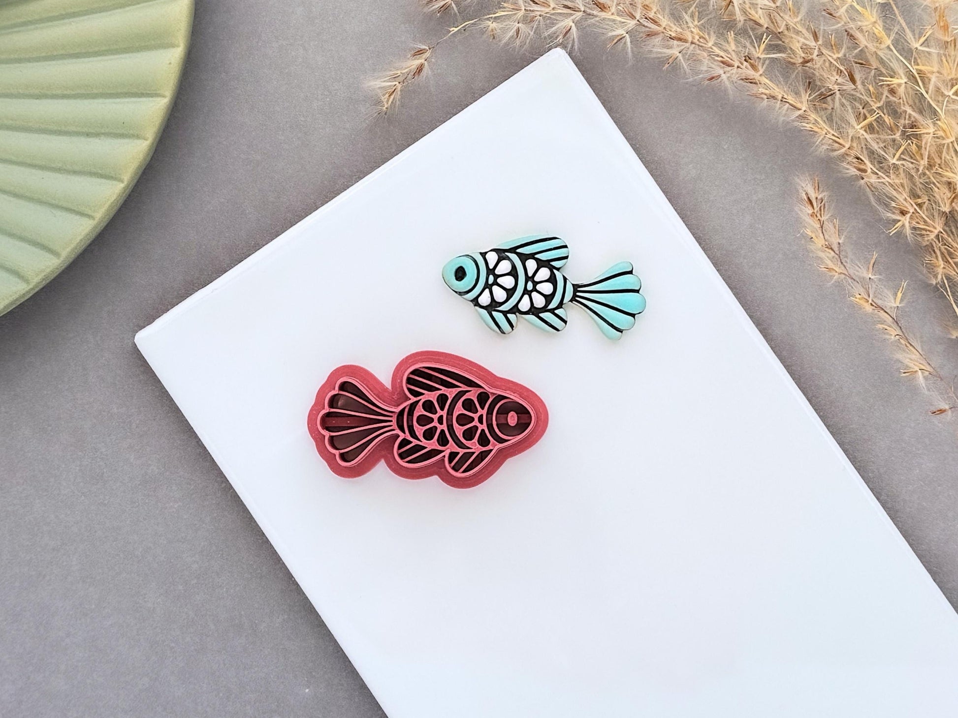 Ornamental fish polymer clay cutter displayed beside a matching pale teal clay cut-out. The folk art-inspired fish design features decorative scales and flowing fins and is photographed on a white ceramic tile.